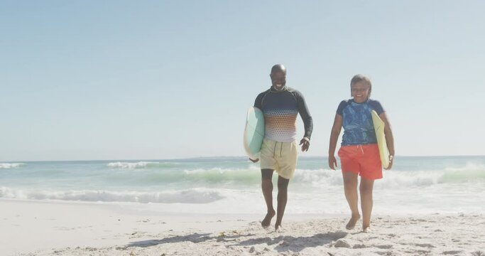 Happy senior african american couple walking with surfboards on sunny beach