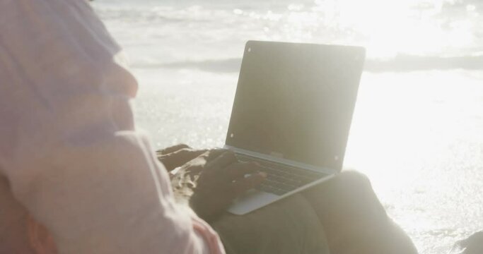Senior african american man sitting and using laptop on sunny beach