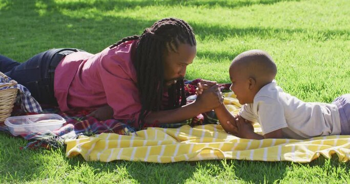 Video of happy african american father and son having picnic on grass, arm wrestling