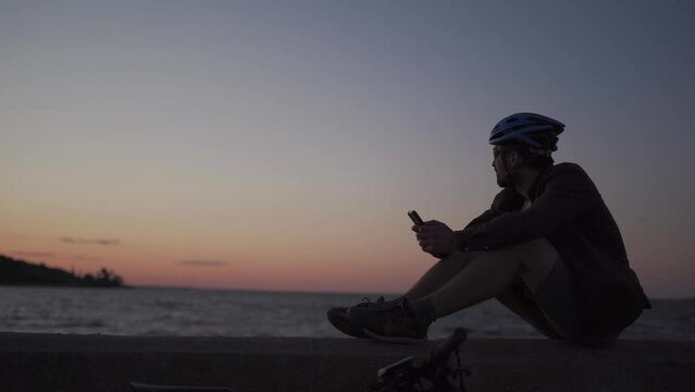 Healthy, active lifestyle and cycling travel. Caucasian man rode bicycle to sea and stopped to plot route on smartphone navigator. Cyclist at sunset by the sea uses a smart phone in windy weather.