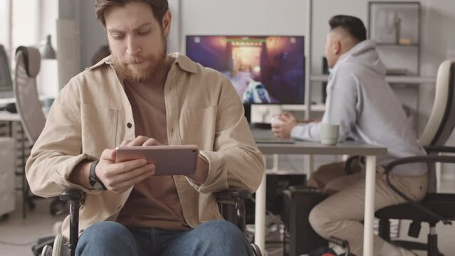 Tilt up slowmo portrait of Caucasian video game developer with tablet PC sitting in wheelchair at modern programmers office smiling at camera with fps game on computer monitor in background