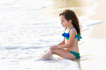 A little girl 6 years old in a mermaid swimsuit sits on a sea rocky coast. The wind blows hair and...