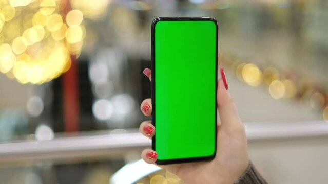 female hand holding smartphone with blank green screen chroma key walks in the mall. Young wife on the background of New Year's decorations in a shopping center. girl holding a smartphone with a green