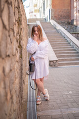 Beautiful and sexy girl in a white dress and white sandals on a high platform blouse with big breasts and a deep neckline posing against a wall on the streets of the city