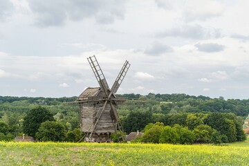 An old wooden mill in the middle of a green field near the houses in the village against the backdrop of a cloudy sky. Ukraine. Pirogovo