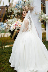 Fototapeta na wymiar Full length portrait of attractive young bride in a white wedding dress and veil holding a big bouquet of white and pink flowers in her hands on a background of wedding ceremony arch