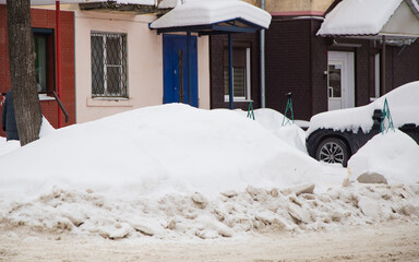 A loose snowdrift by the road against the backdrop of a city street with cars. On the road lies dirty snow in high heaps. Urban winter landscape. Cloudy winter day, soft light.