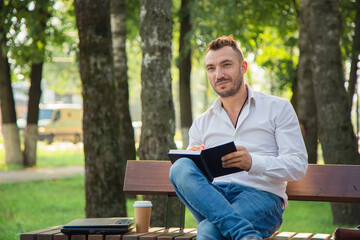 A smiling man sits on a bench, makes plans, writes in a notebook. A young man on a background of green trees, a hot sunny summer day. Warm soft light, close-up.
