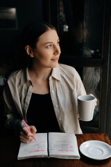 A young European freelancer girl with long dark hair in a shirt remotely works in a cafe, businesswoman are writing notes with pink pen, drinking black coffee, smiling and looking on the street