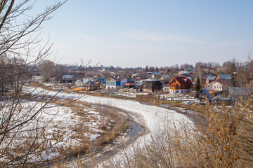 A curved, frozen river stretching into the distance, a village on the shore. Spring, snow melts, puddles and dry grass all around. Day, cloudy weather, soft warm light.