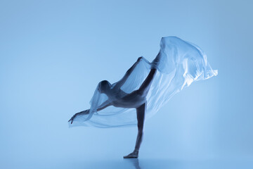 Portrait of flexible woman, graceful ballerina dancing with fabric, cloth isolated on blue studio background. Grace, art, beauty concept. Weightless, tenderness.