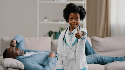 Little african american daughter playing with dad father patient lying on couch kid girl pretending to be doctor looking at camera smiling showing thumb up no problem excellent health approval gesture - Powered by Adobe
