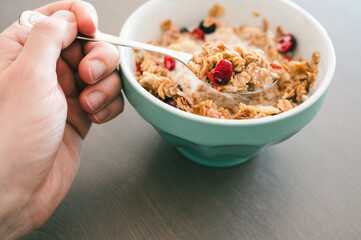 Young man's hand with muesli bowl, using spoon for eating cereals with fruits and milk as a...