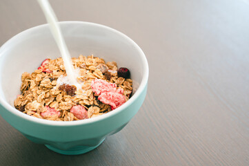 Milk pouring into bowl of fresh muesli, side view with copy space,  isolated on wooden background. Healthy breakfast concept.