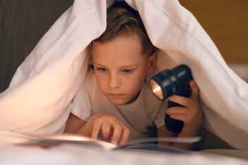 Caucasian child boy reading interesting book under the covers at night
