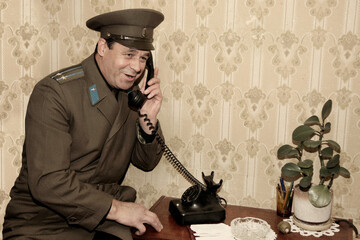 Portrait of a man in the form of a Soviet officer with a telephone receiver, talking on an old landline phone. - obrazy, fototapety, plakaty