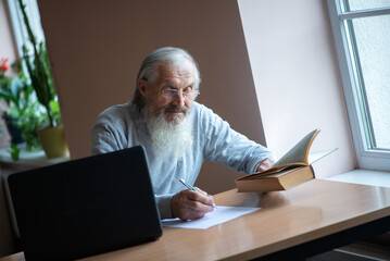 Bearded old man with laptop and book sitting at table and write notes