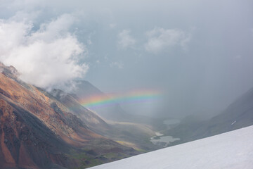 Alpine landscape with vivid rainbow above mountain lake. Scenic view from snow hill to bright rainbow above lake in mountain valley. Top view from snow mountain to colorful rainbow and low clouds.