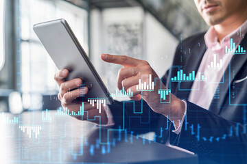 Businessman in suit using tablet device to optimize trading strategy at corporate finance fund. Forex chart hologram over modern panoramic office background