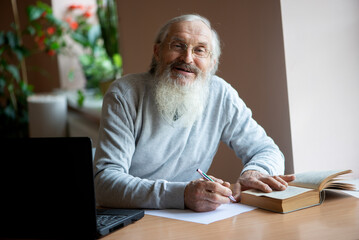 Bearded old man with laptop and book sitting at table and write notes.. © Zelma