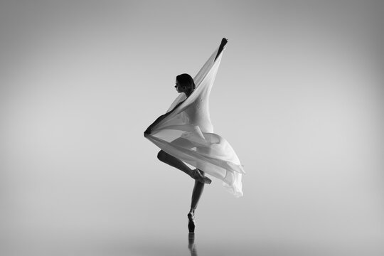 Black and white portrait of graceful ballerina dancing with fabric, cloth isolated on grey studio background. Grace, art, beauty concept. Weightless, flexible.