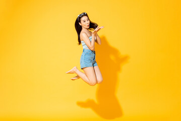 Summer portrait of young Asian woman jumping and blowing kiss in isolated studio yellow color...