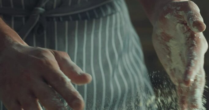Cinematic macro shot of professional artisan baker chef clapping his hands in dust with flour powder while kneading dough for preparation of pasta, pizza and other pastries in rustic bakery kitchen.