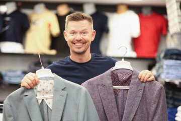 Smiling man is holding two suits on hangers in store