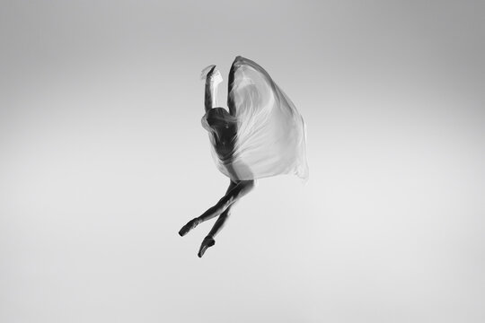 Black and white portrait of graceful ballerina dancing with fabric, cloth isolated on grey studio background. Grace, art, beauty concept. Weightless, flexible.