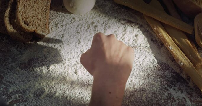 Cinematic close up shot of professional artisan baker chef is drawing heart shape with flour on wooden board with bread, loaf and breadsticks on background in rustic bakery kitchen.