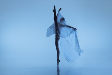 Portrait of flexible woman, graceful ballerina dancing with fabric, cloth isolated on blue studio background. Grace, art, beauty concept. Weightless, tenderness.