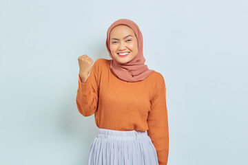 Excited young Asian Muslim woman in brown sweater standing doing winning gesture celebrating fist saying yes isolated over white background. Muslim lifestyle concept