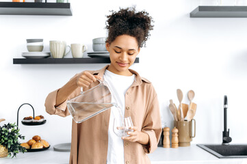 Happy healthy african american girl in casual clothes, standing at home in the kitchen, pouring...