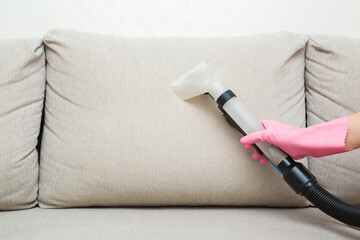 Woman hand in rubber protective glove using spray nozzle of professional vacuum cleaner and washing light beige back pillow of sofa at home. Extraction method. Commercial cleaning service. Closeup.