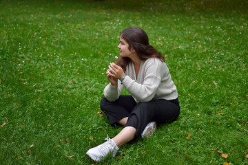 Outdoors, a woman in a beige knitted sweater and trousers sits on the grass in the park, holding a chamomile flower in her hand and looking away, resting and recuperating after a hard day's work.