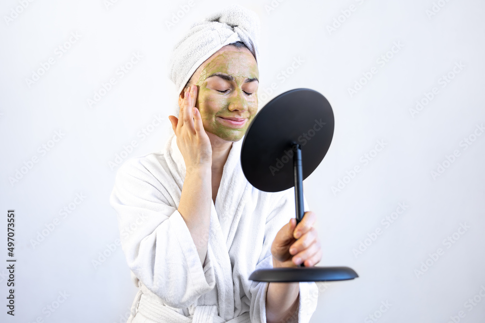 Wall mural young woman with a green mask on her face in a bathrobe in front of a mirror.