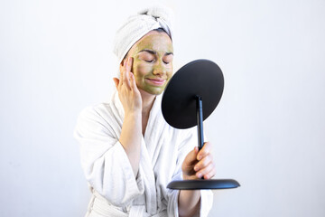 Young woman with a green mask on her face in a bathrobe in front of a mirror.