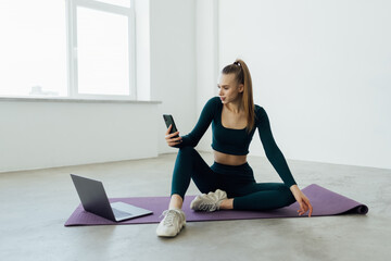 Young woman in sportswear sitting floor resting while doing fitness yoga at home using phone chatting. Losing weight at home. Online class with the laptop concept.