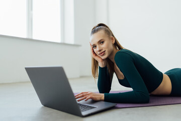 Training Concept. Smiling fit young woman in sports clothes watching fitness videos on internet using laptop near window, lying on floor yoga mat in the gym