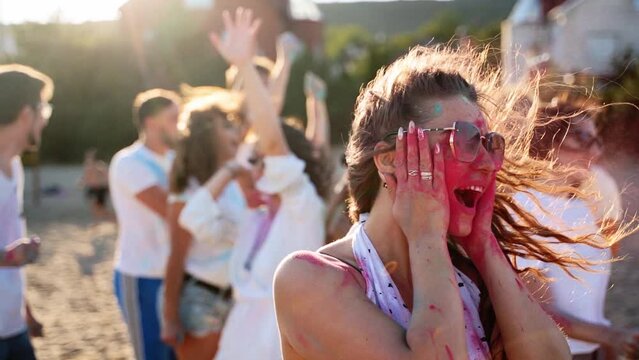 Joyful woman splashes colored powder on cheeks, puts out tongue, has fun at Holi festival in slow motion. People celebrate hindu holiday and end of coronavirus pandemic, quarantine, restrictions