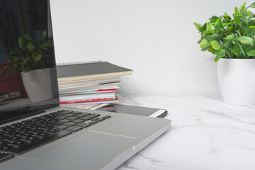 Laptop on white table with potted plant, notebooks and smartphone.