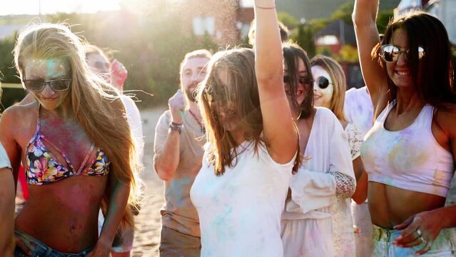 Cheerful people celebrate Holi festival stained in colored powder. Friends have fun at hindu holiday of spring, colors, love. Guys dance, jump on beach in slow motion. End of covid pandemic isolation.