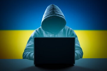 Faceless hooded anonymous computer hacker on Ukrainian flag background. Internet crime and electronic banking security. Anonymous person in the hood sitting in front of computer working with laptop