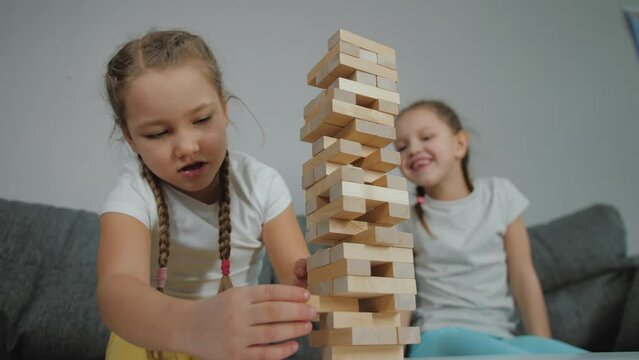 Funny cute caucasian sisters playing building blocks together at home. Little girl takes wooden brick, tower falls and breaks. Table top, board game, Children playing jenga