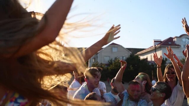 Joyful people stained in colored powder toss guy up in air at Holi festival on beach in slow motion. Friends have fun at hindu holiday of spring, colors, love. End of covid pandemic isolation.