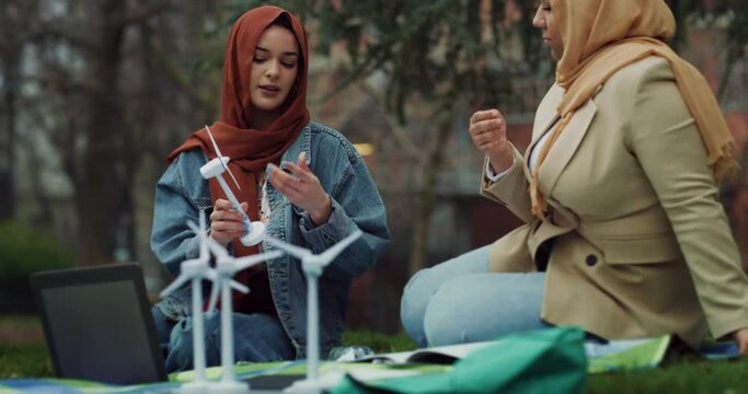 Cinematic shot of young happy Arabian muslim female students friends wearing hijab work together on university engineering project of wind mill farm sustainable green energy in public city park.  