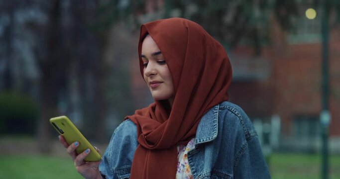 Cinematic shot of young happy Arabian muslim woman wearing hijab is having fun to use smartphone for navigate in internet and comunicate with friends on social media in green city park.