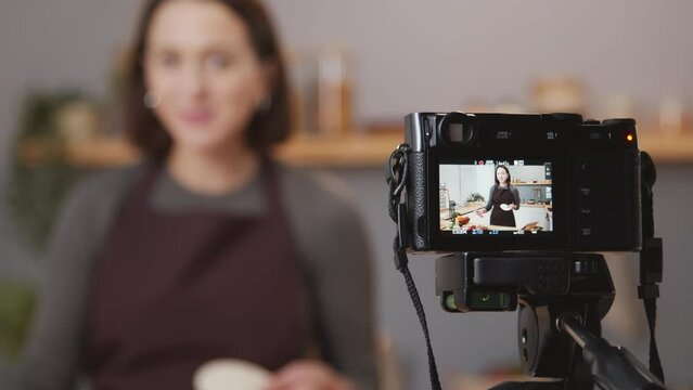 Selective focus shot of professional camera on tripod filming female food blogger telling dinner recipe in kitchen