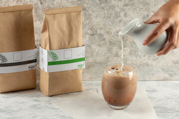 A hand pouring milk over a glass with coffee and two bags of coffee with a label