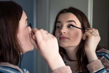 A young woman paints her eyelashes with mascara in front of a mirror.